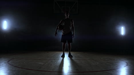 Basketball-player-in-sportswear-red-shorts-and-a-blue-t-shirt-goes-on-a-dark-basketball-court-in-the-backlighting-coming-out-of-the-smoke-knocks-a-basketball-ball-on-the-floor-looking-at-the-camera-in-slow-motion